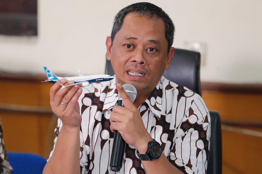 A man is holding up a model of a plane in one hand while holding a microphone to his mouth in the other