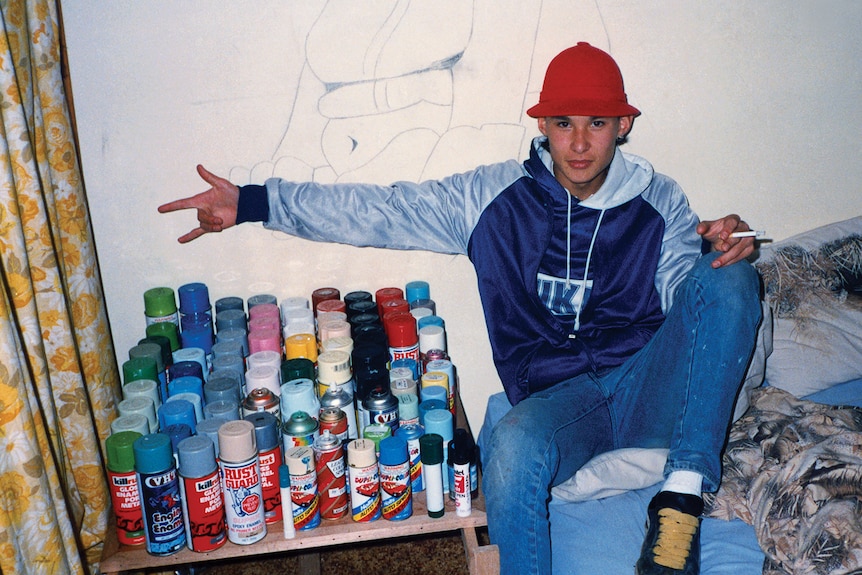 A teenager holding a cigarette points to a large collection of spray cans