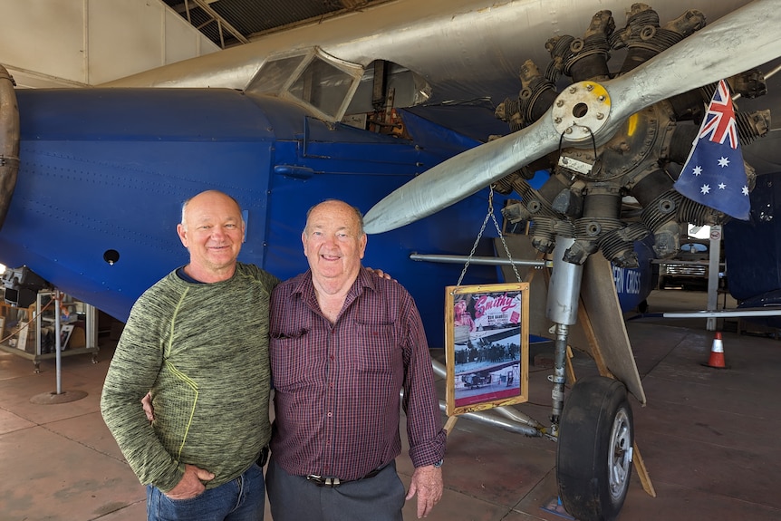 Two men  stand in front of a replica light aeroplane
