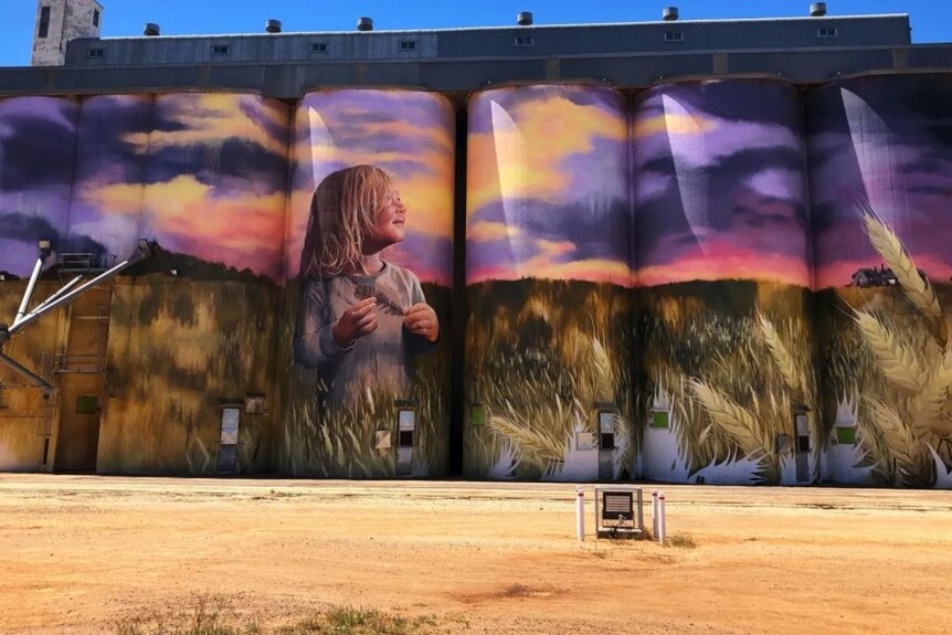 A line of art silos with a mural of a little girl standing in a wheat field against a beautiful sunset