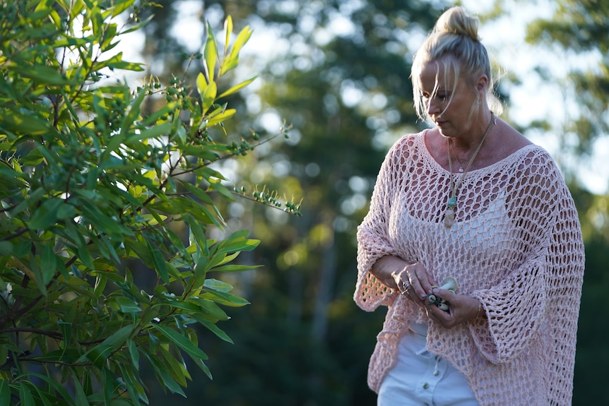 A woman in a blush pink shawl and white pants stands in a garden looking down at the ground