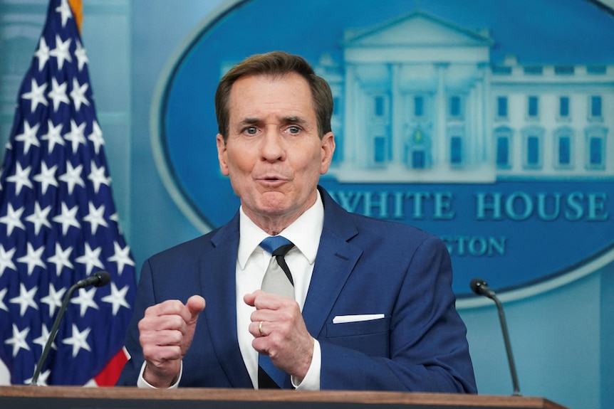 A white man in a suit and tie stands and speaks at a lecturn, clenching his hands. American flag and White House image behind