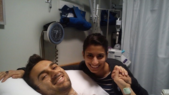 Young man in hospital bed holding hands with a young woman who's seated at his bedside