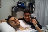 Young man in hospital bed holding hands with a young woman who's seated at his bedside