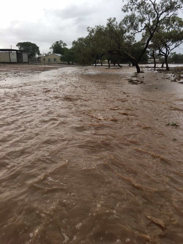 Flooding covers a property with brown water, in the distance there are trees and buildings.