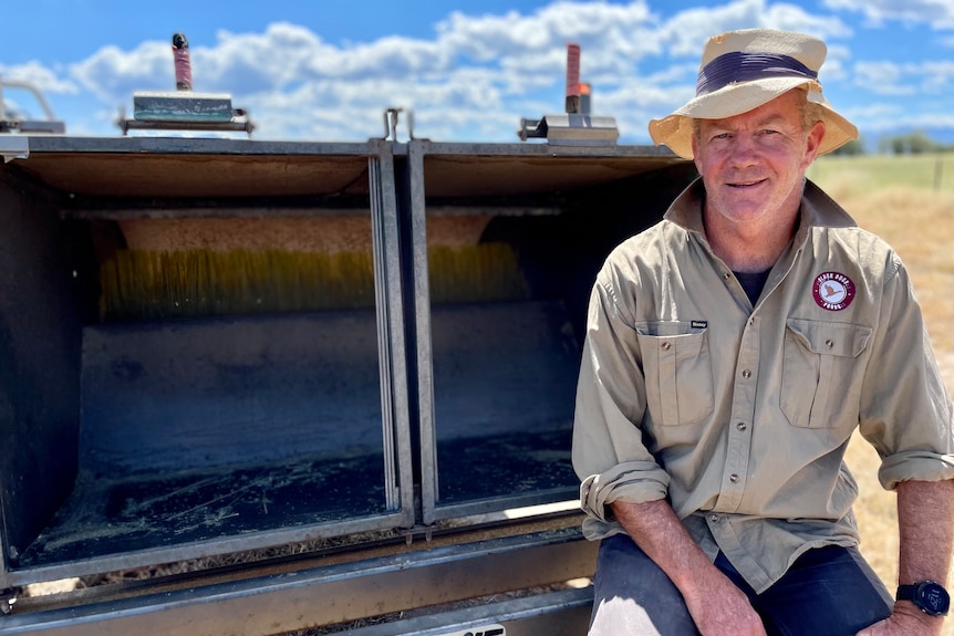 A man in a hat and khaki shirt sits in front of a small harvester.