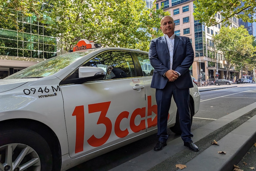 A bald man wearing a suit poses next to a 13cabs cab on a leafy street.