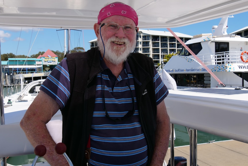 An older man stands on a sailing boat in a blue and navy striped shirt, jacket, reading glasses and pink bandana