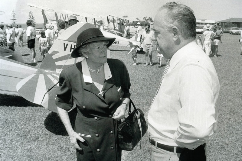 An older woman and man, dressed in smart clothing, stand at an airfield with light aircraft behind them.