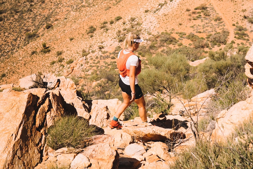 Runner wearing a white t-shirt and black exercise pants and wearing running vest on rocky hill.