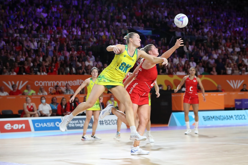 An English netball goal shooter reaches out to grab the ball as an Australian goal keeper tries to spoil from behind.