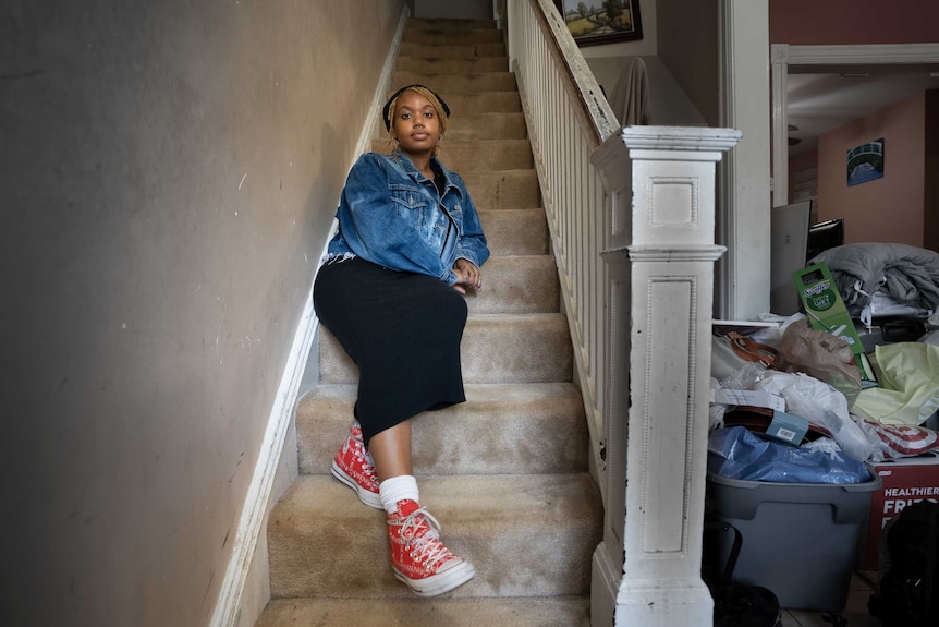 A young woman sits on the staircase inside her house looking solemn