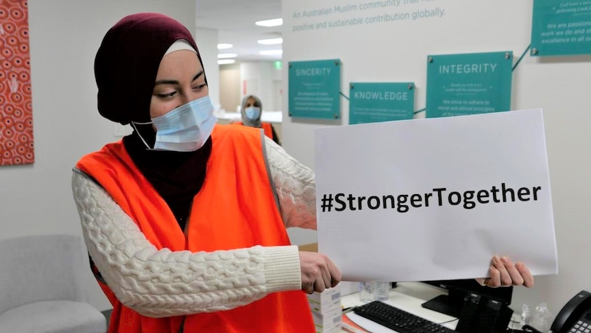 A woman in a hijab and hi-vis vest holds a sign reading "Stronger Together".
