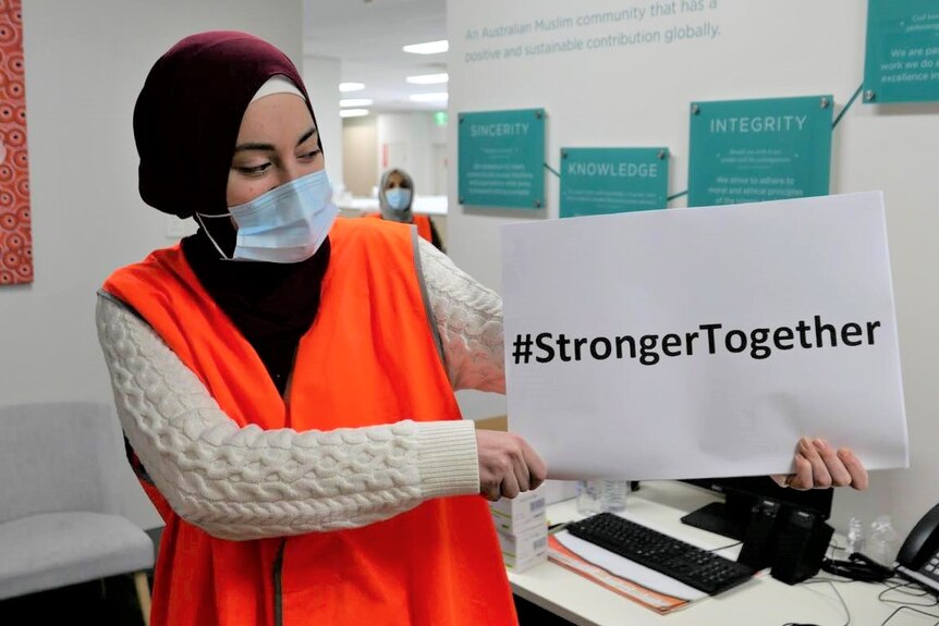 A woman in a hijab and hi-vis vest holds a sign reading "Stronger Together".