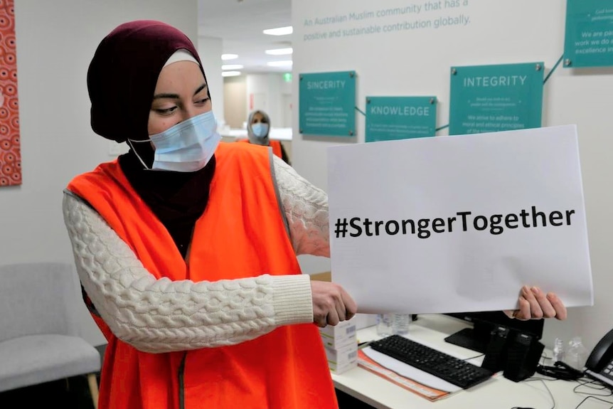 A woman in a hijab and hi-vis vest holds a sign reading "Stronger Together".