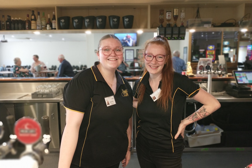Two girls stand behind a bar smiling at the camera with patrons in the background.