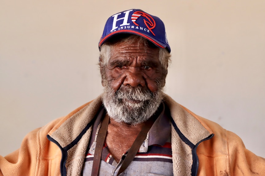 An aboriginal elder with a grey beard wearing a blue baseball cap and an orange jumper 