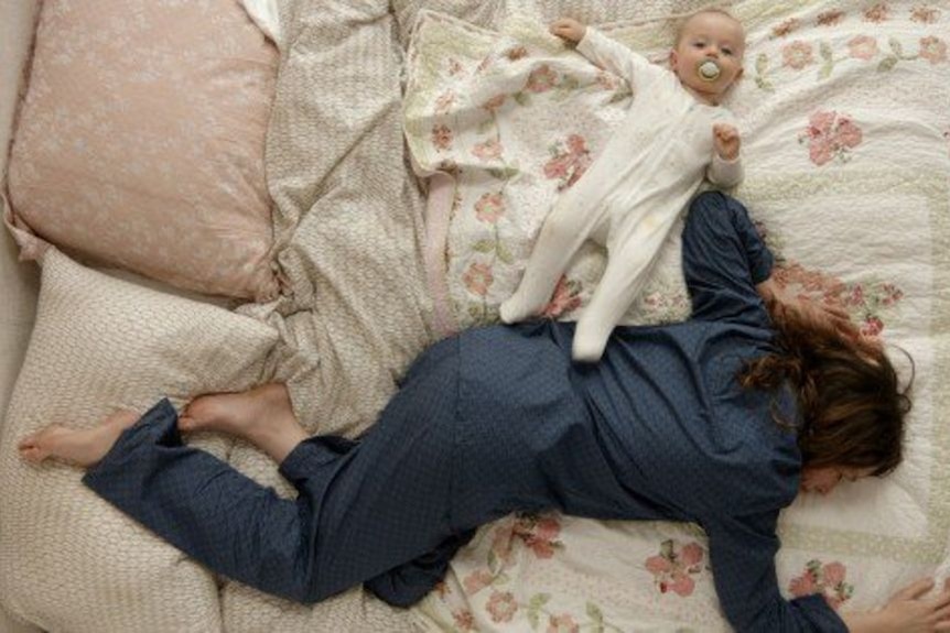 A still image from the ABC comedy show The Letdown, showing actor Alison Bell lying on the bed next to a baby.