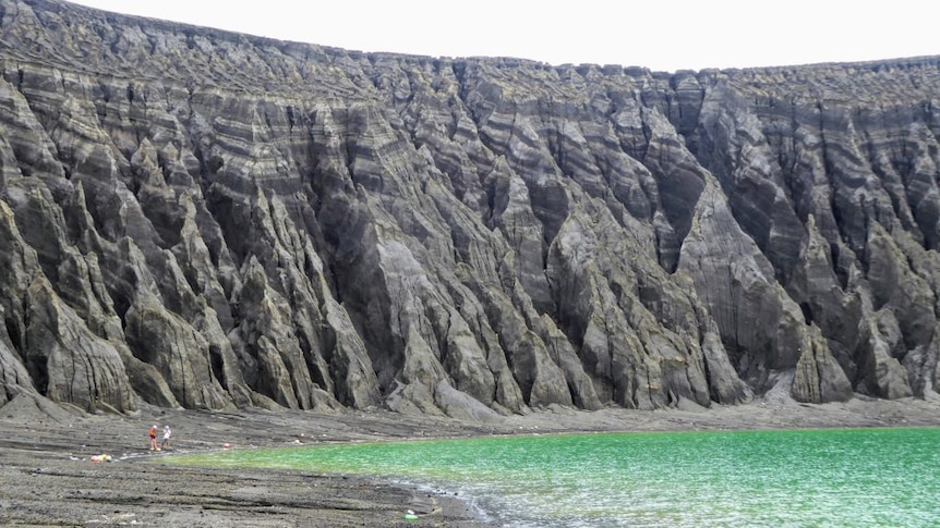 A blue-green crater lake in the foreground with a volcanic rim in the background lined with eroded gullies
