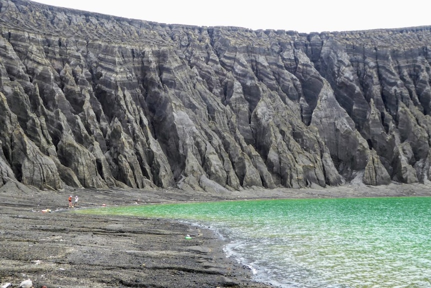 A blue-green crater lake in the foreground with a volcanic rim in the background lined with eroded gullies
