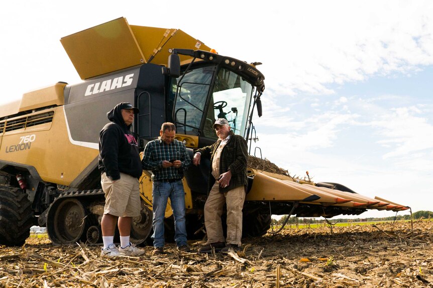 Three men lean against a  yellow corn harvester.