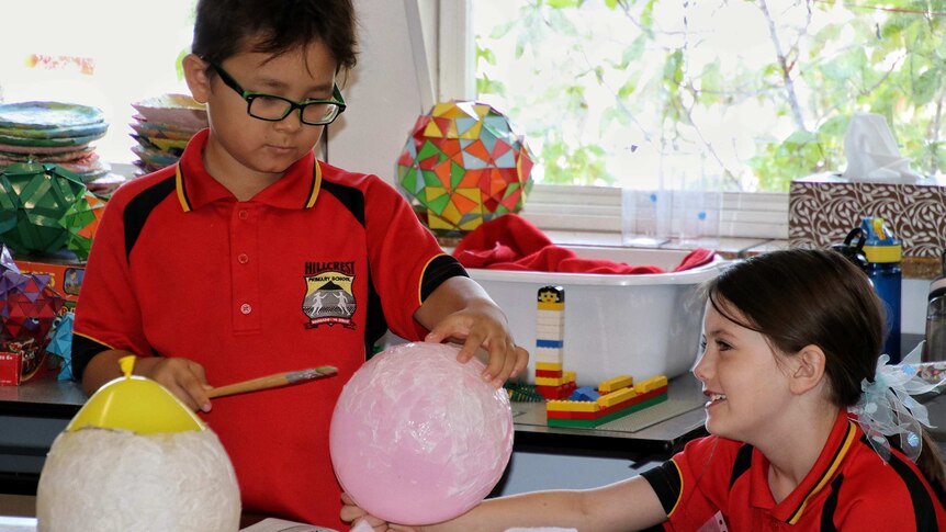 Two primary school students in red shirts creating paper lanterns at a school desk.