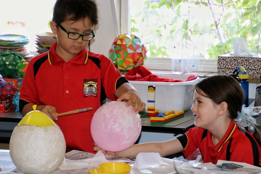 Two primary school students in red shirts creating paper lanterns at a school desk.