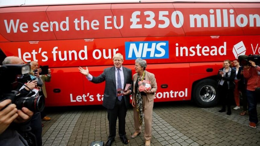 Blonde man in dark suit stands with dark grey haired woman in beige suit in front of red bus