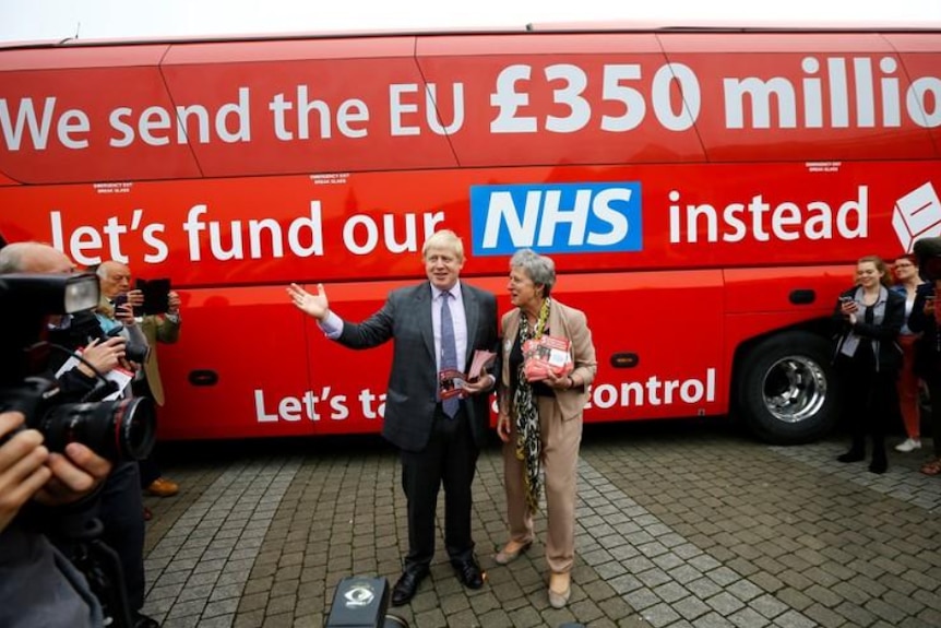 Blonde man in dark suit stands with dark grey haired woman in beige suit in front of red bus