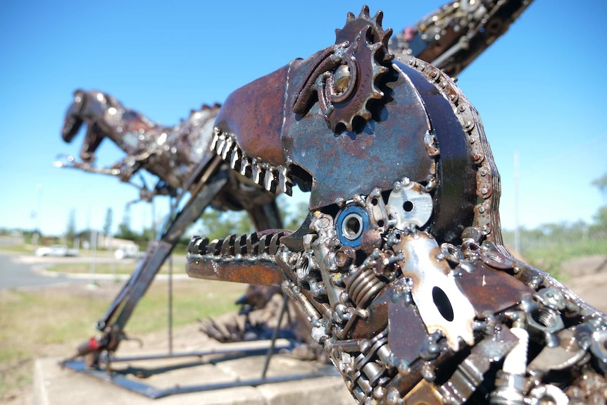 Close up of scrap metal dinosaur neck and head, different parts welded together with larger dinosaur in the background.