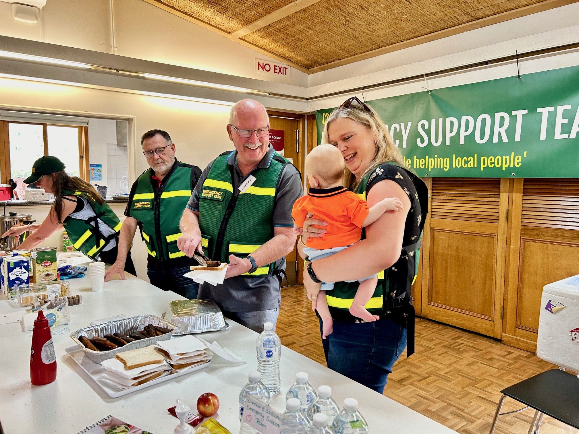 A woman holding a baby talks to a man who is preparing food.