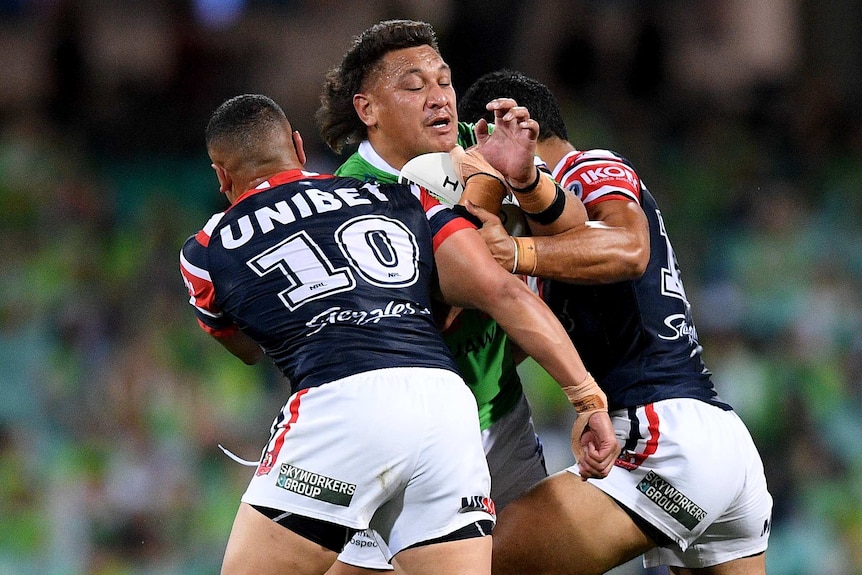 A Canberra Raiders NRL player is tackled across his upper body by two Sydney Roosters opponents.