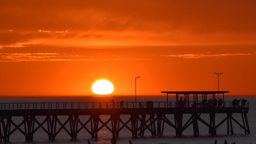 People walk along Largs Jetty at Largs Bay in Adelaide as the sun starts to dip below the horizon.