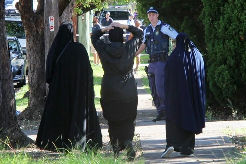 Three women in hijabs speak to a policeman standing in front of a house