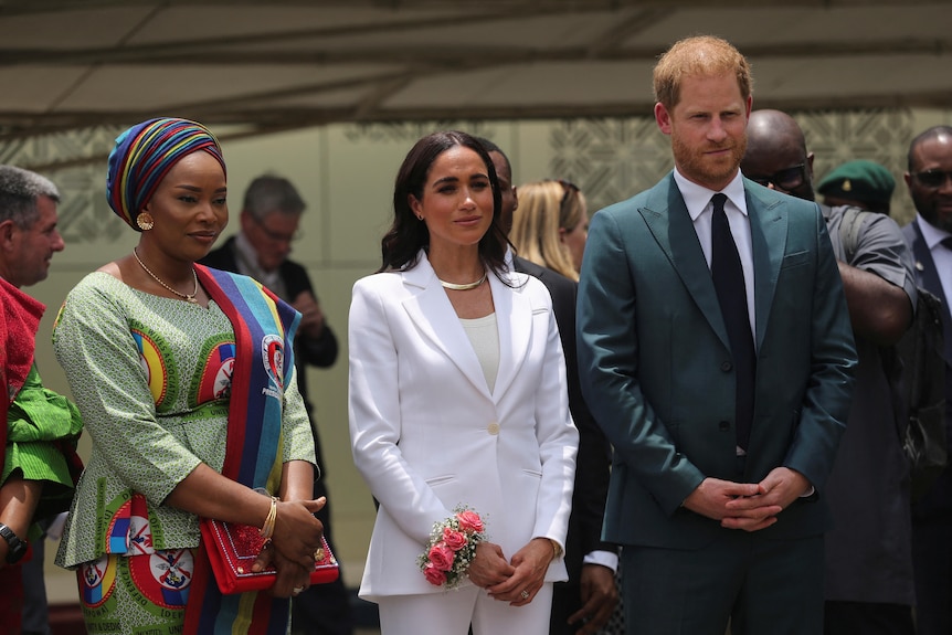 A woman wearing a rainbow scarf on her head and green dress stands next to Meghan and Prince Harry.
