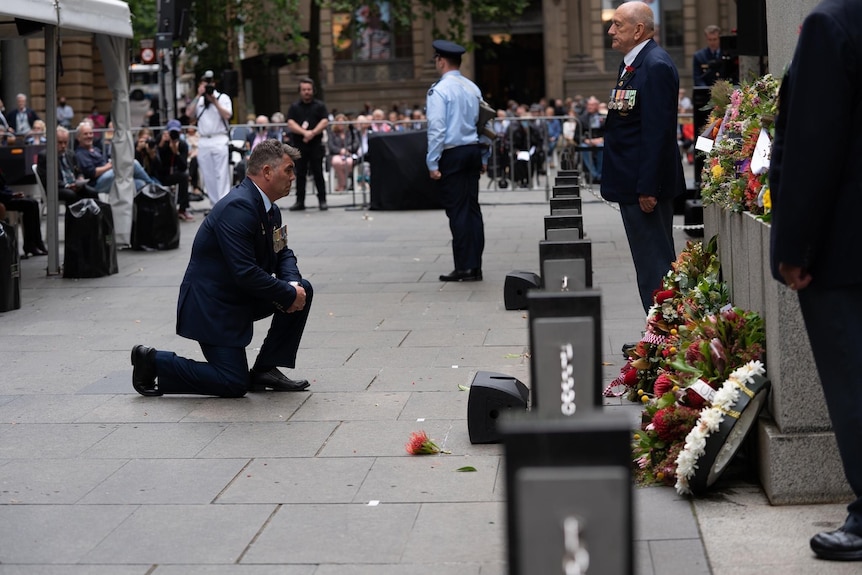 A man wearing a suit kneeling on the pavement in front of a war memorial that is covered in floral wreaths.