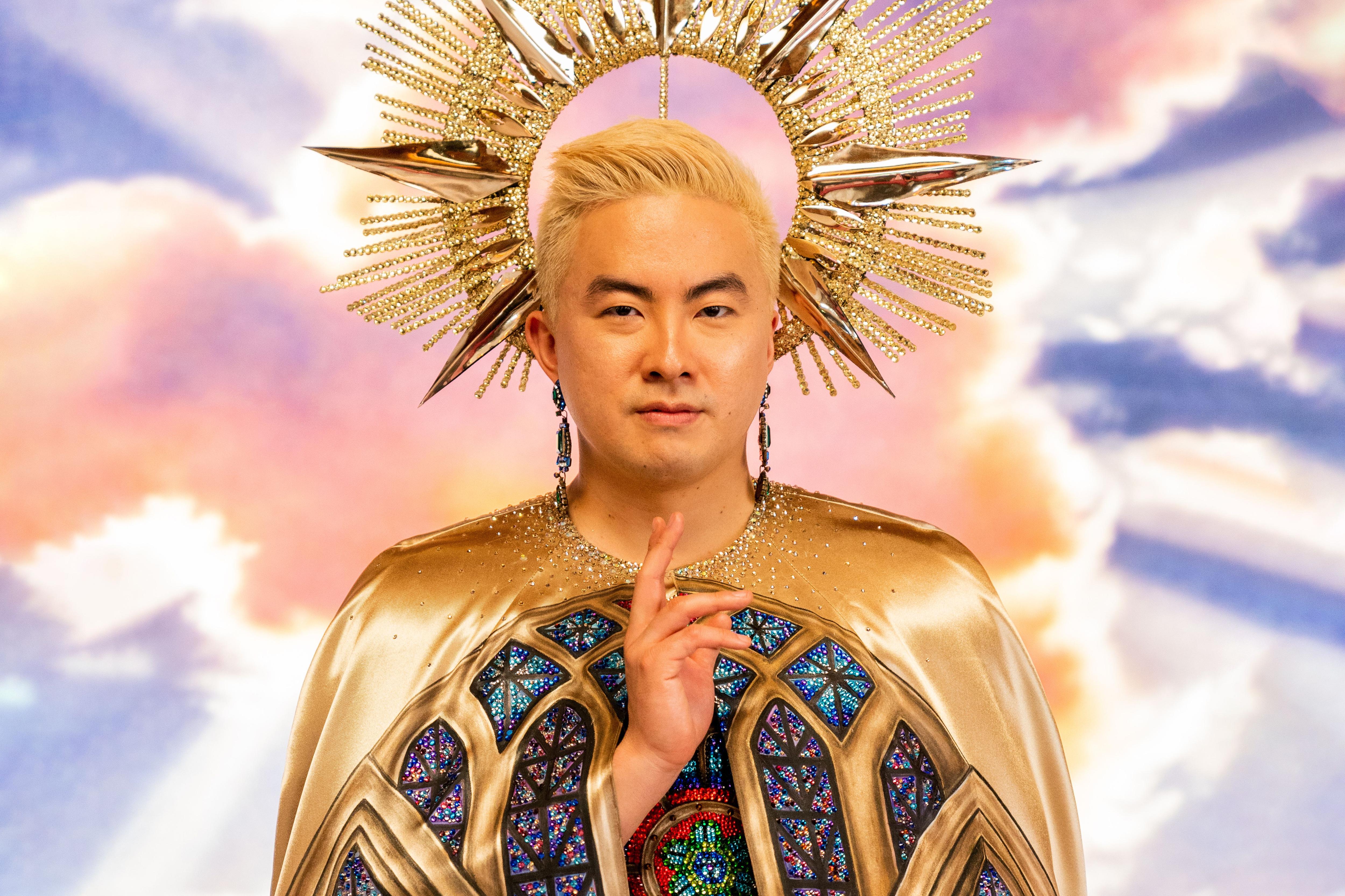 Bowen Yang looks at the camera, wearing an elaborate gold headdress and gold cape, against a backdrop of heavenly clouds.