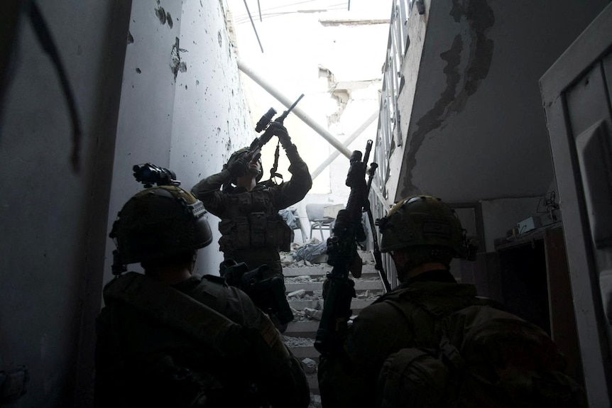 A solldier peers along the barrel of their rifle up a stairwell in a wrecked, bullet-ridden home.