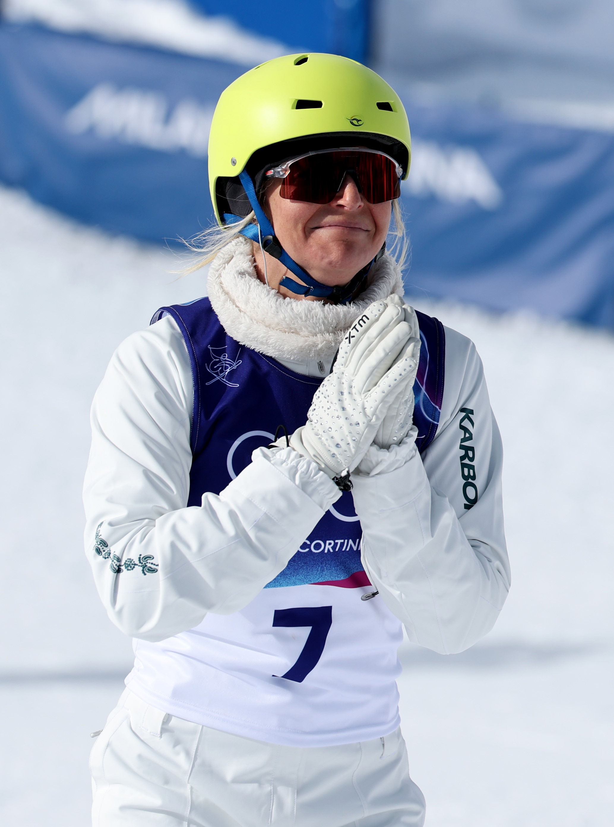 Uma mulher loira com capacete e equipamento de esqui junta as mãos em um campo de neve.