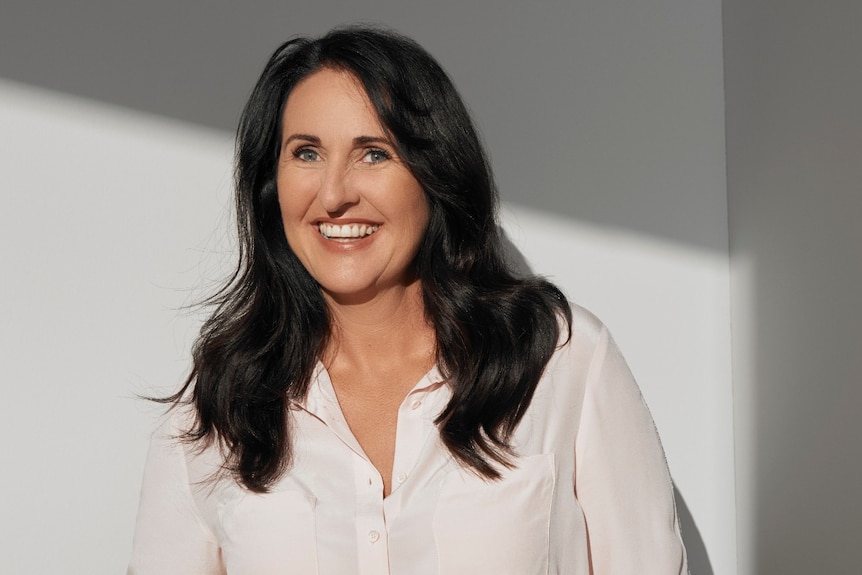 A woman with dark hair against a plain wall smiles at the camera