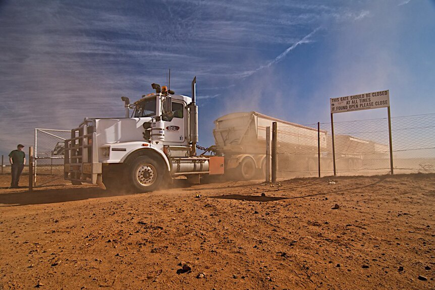 A white road train passing through a gate on a red dirt road