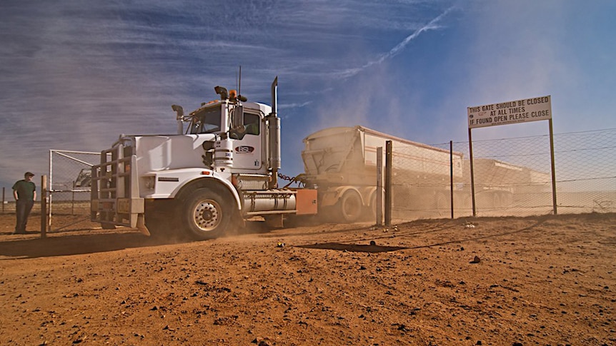 A white road train passing through a gate on a red dirt road