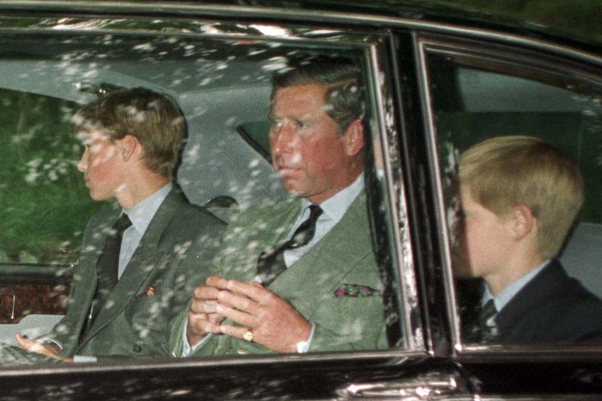 Prince Charles with sons William (15) and Harry (11) in the back of a car, looking away from the camera
