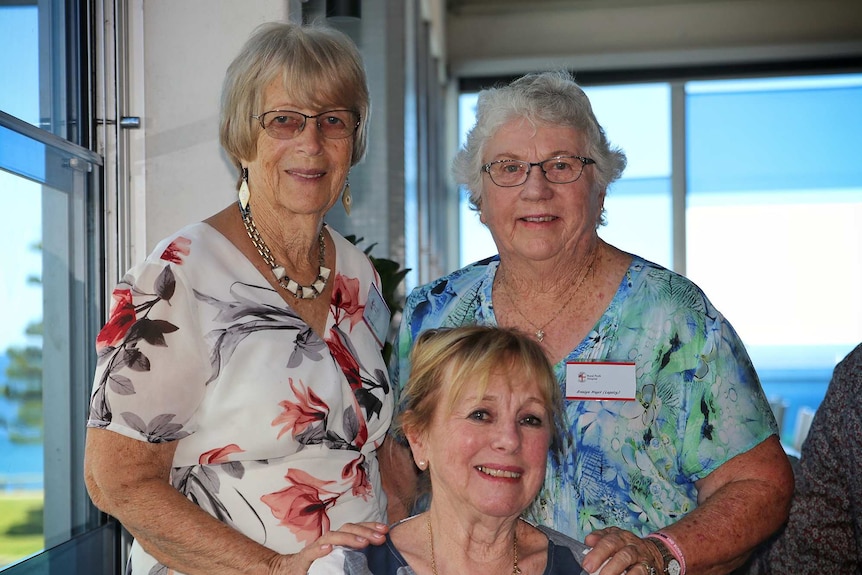 Three older women pose smiling for a photo with two of them standing up behind the third.