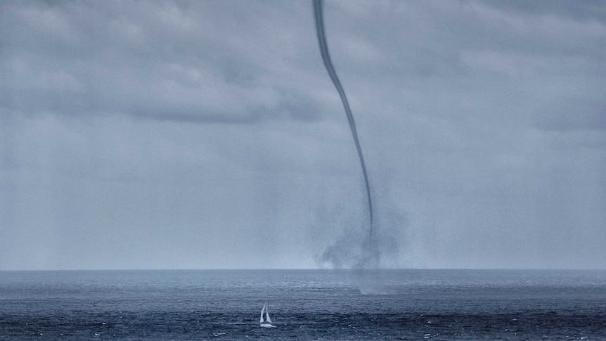 A yacht sails past a water spout reaches down from a cloud and touches down on the ocean.