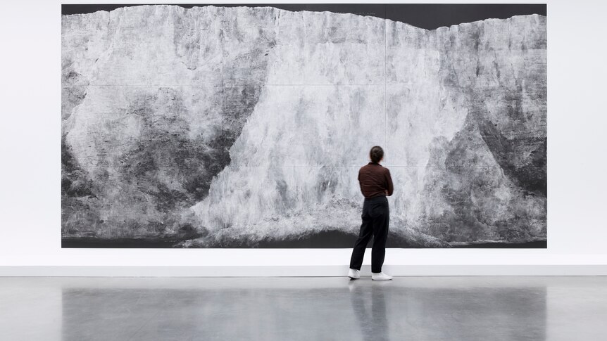 A photograph of a person standing in front of a large chalk illustration hanging in a gallery depicting a crumbling chalk cliff