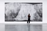 A photograph of a person standing in front of a large chalk illustration hanging in a gallery depicting a crumbling chalk cliff