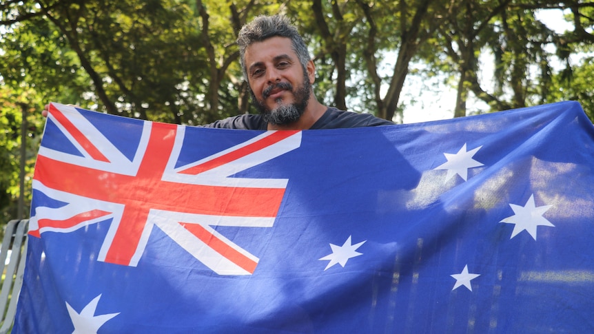 A man standing, holding the Australian flag outstretched and smiling, in front of trees and blue sky.