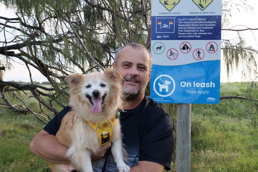 A man in his 40s holding a pomeranian dog in front of an 'on leash' sign. 