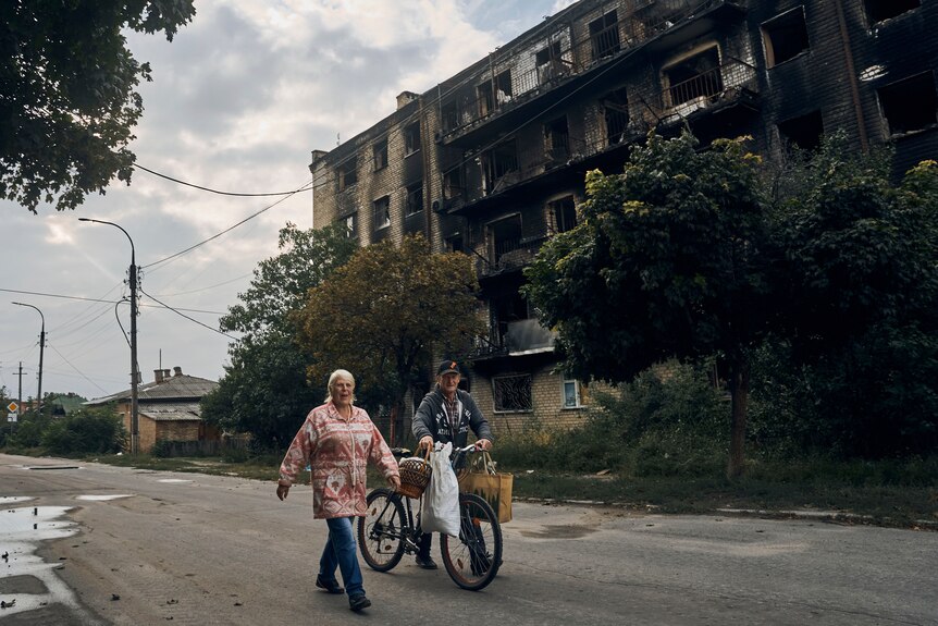 Une femme dans une veste lumineuse et un homme poussant un vélo marchent sur une route bordée de bâtiments endommagés